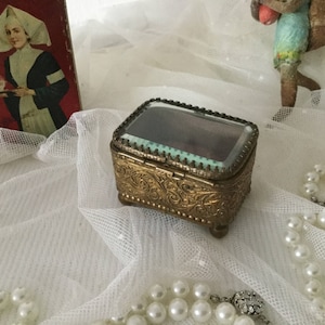 May include: A small, antique brass jewelry box with a glass top, ornate floral detailing, and small feet. A vintage advertisement and a strand of faux pearls are in the background. The box is rectangular and has a hinged lid.
