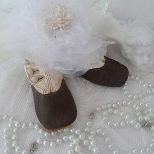 May include: A pair of brown leather baby shoes with white lace and pearl accents. The shoes are on a white lace tablecloth with a pearl necklace.