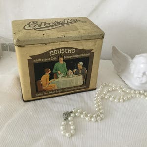 Könnte beinhalten: Eine vintage Blechdose mit cremefarbenem Hintergrund und braunen Schriftzügen. Die Dose zeigt ein Logo mit dem Text "EduScho" und den Worten "schafft zu jeder Zeit ein Behagen u Gemütlichkeit". Die Dose hat ein Bild von drei Frauen, die an einem Tisch mit einer weißen Tischdecke sitzen. Die Dose steht auf einer weißen Oberfläche.