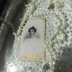 May include: A vintage black and white photograph of a woman wearing a white blouse and a pearl necklace. The photo is framed and placed on a white lace tablecloth.