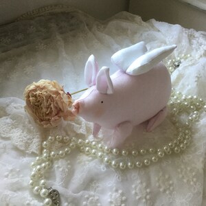 K&ouml;nnte beinhalten: Ein rosa Schwein mit wei&szlig;en Fl&uuml;geln, das auf einem wei&szlig;en Spitzenstoff mit einer Perlenkette und einer getrockneten Blume sitzt.