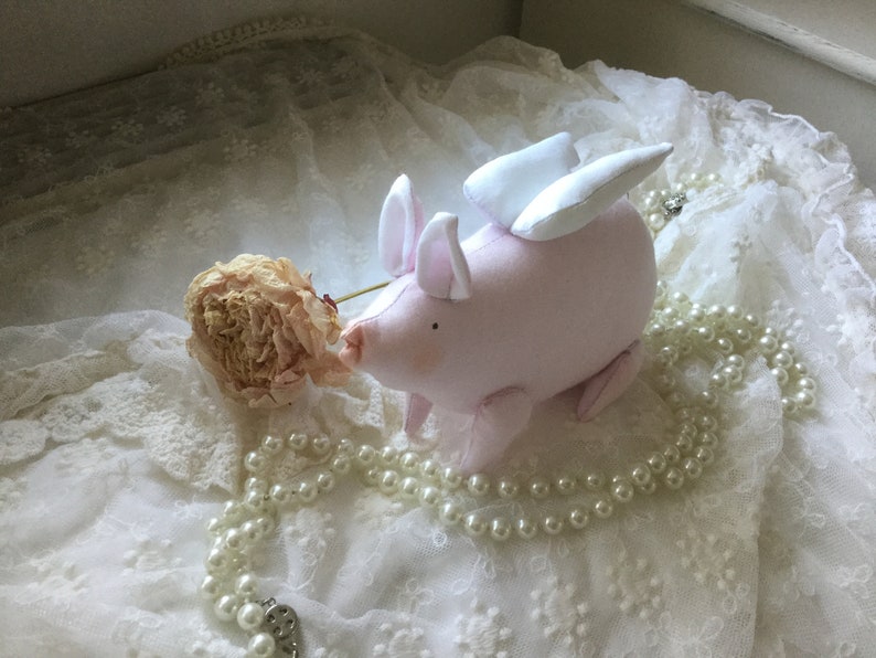 K&ouml;nnte beinhalten: Ein rosa Schwein mit wei&szlig;en Fl&uuml;geln, das auf einem wei&szlig;en Spitzenstoff mit einer Perlenkette und einer getrockneten Blume sitzt.