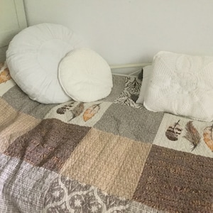 May include: A white bedspread with a patchwork design featuring brown, beige, and gray squares. The bedspread has a pattern of feathers in the center. Three white pillows are on the bedspread.
