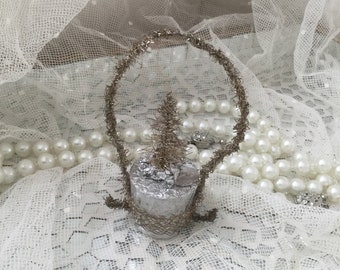 Könnte beinhalten: Ein silberner und brauner Weihnachtsbaumschmuck aus Draht mit einer silbernen Basis und einem Drahtrahmen. Der Schmuck liegt auf einer weißen Spitzentischdecke.