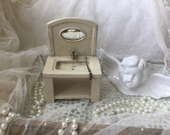 Könnte beinhalten: Ein weißes Miniatur-Badwaschbecken für ein Puppenhaus mit einem Spiegel, einem Wasserhahn und einer Kette, die vom Wasserhahn hängt. Das Waschbecken steht auf einem weißen Holzgestell. Das Waschbecken ist umgeben von weißen Perlen und einer weißen Porzellan-Engelfigur.