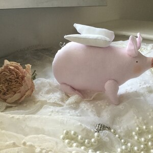K&ouml;nnte beinhalten: Ein rosa Stoffschwein mit wei&szlig;en Fl&uuml;geln, das auf einer wei&szlig;en Spitze mit einer Perlenkette sitzt.
