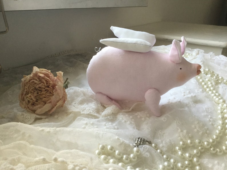 K&ouml;nnte beinhalten: Ein rosa Stoffschwein mit wei&szlig;en Fl&uuml;geln, das auf einer wei&szlig;en Spitze mit einer Perlenkette sitzt.