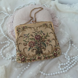 May include: A vintage tapestry clutch purse with a gold chain strap. The purse has a floral pattern with pink, red, and green flowers on a beige background.