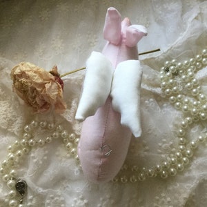 K&ouml;nnte beinhalten: Ein rosa Stoffschwein mit wei&szlig;en Stofffl&uuml;geln. Das Schwein liegt auf einem wei&szlig;en Spitzenstoff mit einer Schnur wei&szlig;er Perlen.