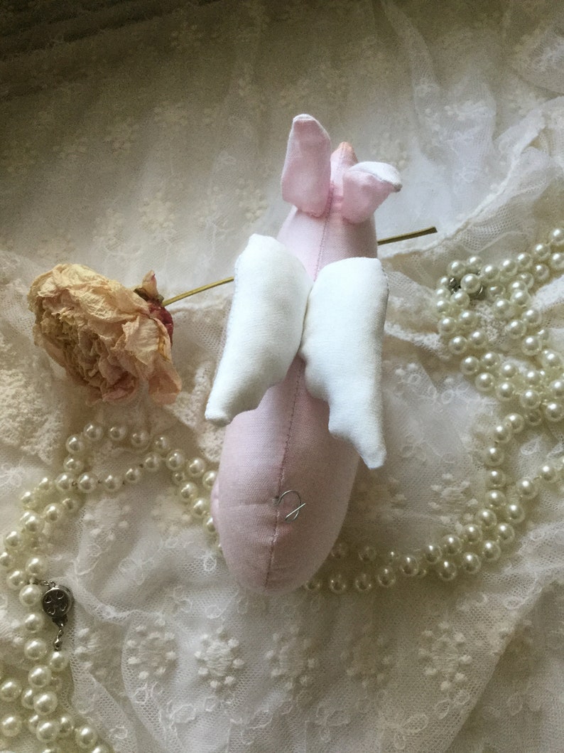 K&ouml;nnte beinhalten: Ein rosa Stoffschwein mit wei&szlig;en Stofffl&uuml;geln. Das Schwein liegt auf einem wei&szlig;en Spitzenstoff mit einer Schnur wei&szlig;er Perlen.