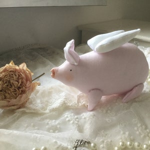 K&ouml;nnte beinhalten: Ein rosa Stoffschwein mit wei&szlig;en Fl&uuml;geln, das auf einer wei&szlig;en Spitze mit einer getrockneten Blume sitzt.