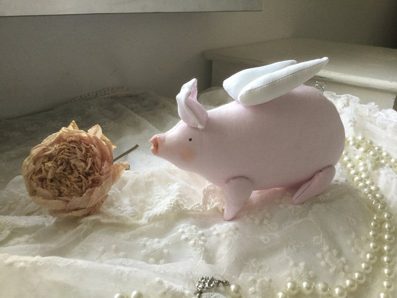 K&ouml;nnte beinhalten: Ein rosa Stoffschwein mit wei&szlig;en Fl&uuml;geln, das auf einer wei&szlig;en Spitze mit einer getrockneten Blume sitzt.