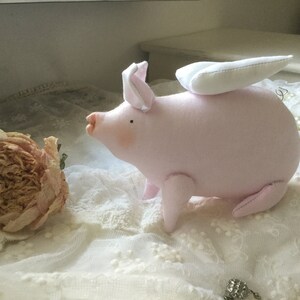 K&ouml;nnte beinhalten: Ein rosa Stoffschwein mit wei&szlig;en Fl&uuml;geln, das auf einer wei&szlig;en Spitze liegt. Das Schwein hat eine kleine, rosa Nase und schwarze Augen.