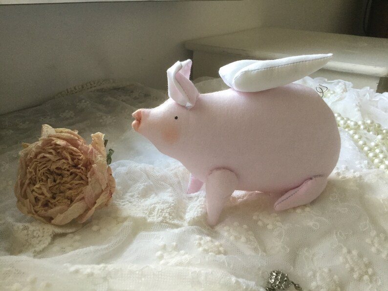 K&ouml;nnte beinhalten: Ein rosa Stoffschwein mit wei&szlig;en Fl&uuml;geln, das auf einer wei&szlig;en Spitze liegt. Das Schwein hat eine kleine, rosa Nase und schwarze Augen.