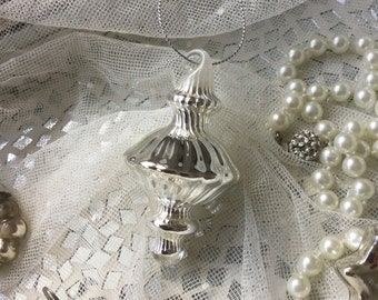 Könnte beinhalten: Ein silberner gläserner Weihnachtsbaumschmuck mit einer spitzen Spitze und einem gerippten Design. Der Schmuck hängt an einer silbernen Schnur und ist von weißer Spitze und Perlen umgeben.