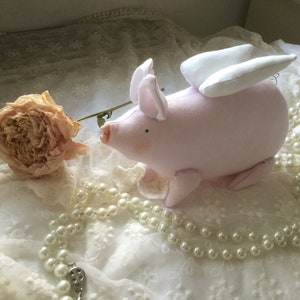 K&ouml;nnte beinhalten: Ein rosa Stoffschwein mit wei&szlig;en Fl&uuml;geln, das auf einer wei&szlig;en Spitze mit einer Perlenkette sitzt.