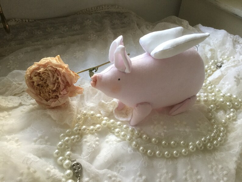 K&ouml;nnte beinhalten: Ein rosa Stoffschwein mit wei&szlig;en Fl&uuml;geln, das auf einer wei&szlig;en Spitze mit einer Perlenkette sitzt.