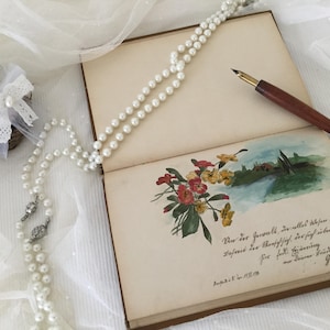 May include: An open book with a pearl necklace and a wooden pen on top. The book has a watercolour painting of flowers and a landscape on the inside page. The text on the page reads: "Un des Inwalt, ür aller Wesen binet, Dachreit der Menschichich, der sich über andet. fire ferli szimering Jeghor"