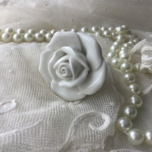 Könnte beinhalten: Eine weiße Porzellanrose mit detailliertem Design, die auf einem weißen Spitzenstoff mit einer Perlenkette liegt.