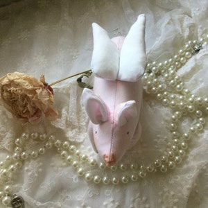 K&ouml;nnte beinhalten: Ein rosa Stoffschwein mit wei&szlig;en Fl&uuml;geln, das auf einer wei&szlig;en Spitze mit einer Perlenkette liegt.
