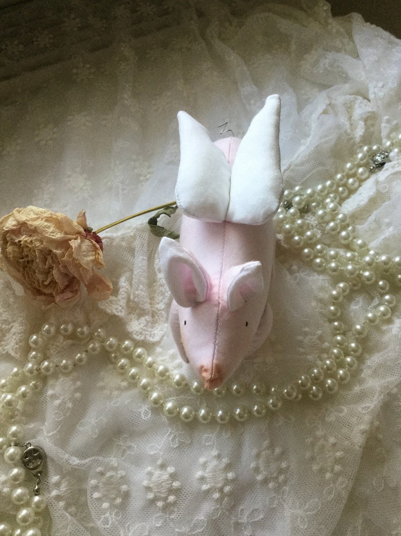 K&ouml;nnte beinhalten: Ein rosa Stoffschwein mit wei&szlig;en Fl&uuml;geln, das auf einer wei&szlig;en Spitze mit einer Perlenkette liegt.