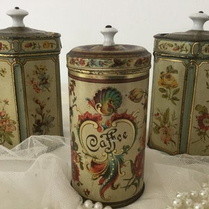 Può includere: Tre barattoli di metallo vintage con disegni floreali. Il barattolo al centro è etichettato "Caffè" e ha un design più elaborato. I barattoli hanno coperchi in porcellana bianca.
