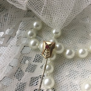 May include: A gold-toned pin with a geometric design and a red gemstone in the centre. The pin is on a white background with a pearl necklace.