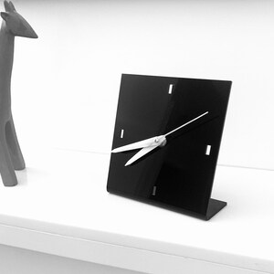 Op de afbeelding: Een moderne zwarte vierkante bureauklok met witte wijzers en een minimalistisch ontwerp. De klok heeft een kleine standaard en staat tegen een witte achtergrond. De wijzers van de klok wijzen naar 10:10.