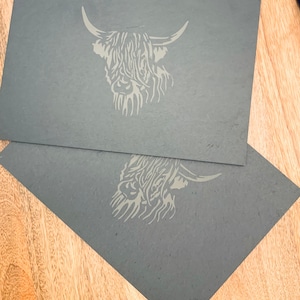 Puede incluir: Dos tarjetas rectangulares grises con una ilustración gris claro de la cabeza de una vaca de las Highlands. La vaca tiene pelo largo y desgreñado y cuernos grandes. Las tarjetas se superponen ligeramente, sobre un fondo de vetas de madera clara.