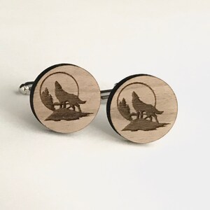 Puede incluir: Un par de gemelos de madera con un diseño de lobo aullando a la luna. Los gemelos son circulares y tienen un acabado de madera marrón claro.