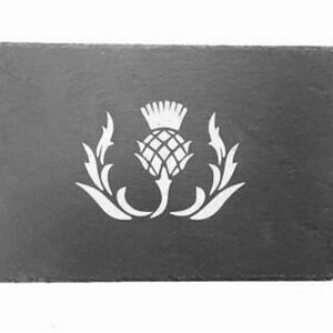 Pode incluir: Um descanso de copo de ardósia preto com uma silhueta branca de um cardo, a flor nacional da Escócia.