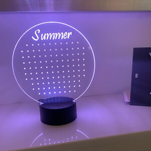 Può includere: Una lampada a LED circolare trasparente con la scritta "Summer" in corsivo. La lampada ha una griglia di piccoli fori distanziati uniformemente e poggia su una base nera. La luce emette un bagliore viola.