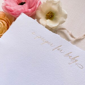 Peut inclure: Une carte blanche avec une inscription dor&eacute;e qui dit "une pri&egrave;re pour b&eacute;b&eacute;". La carte est entour&eacute;e de fleurs roses et blanches.