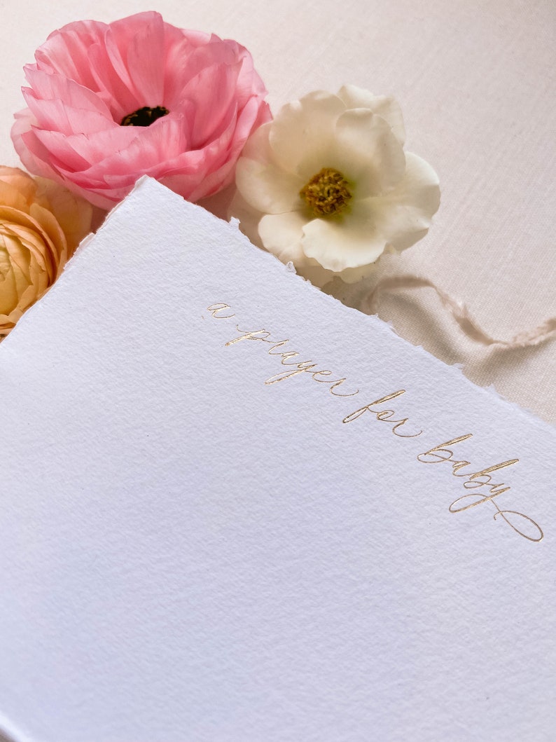 Peut inclure: Une carte blanche avec une inscription dor&eacute;e qui dit "une pri&egrave;re pour b&eacute;b&eacute;". La carte est entour&eacute;e de fleurs roses et blanches.