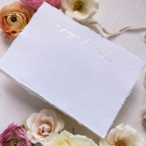 Peut inclure: Une carte blanche avec une inscription dor&eacute;e qui dit "une pri&egrave;re pour b&eacute;b&eacute;". La carte est entour&eacute;e de fleurs roses, blanches et p&ecirc;che.