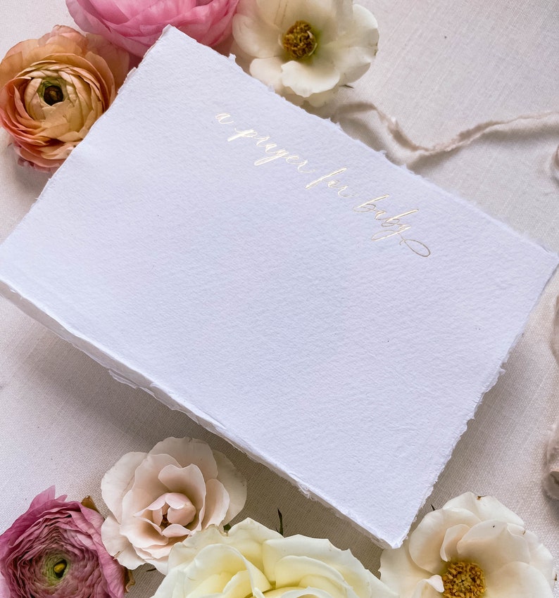 Peut inclure: Une carte blanche avec une inscription dor&eacute;e qui dit "une pri&egrave;re pour b&eacute;b&eacute;". La carte est entour&eacute;e de fleurs roses, blanches et p&ecirc;che.