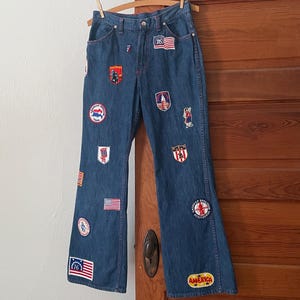 Peut inclure: Jean en denim bleu orné de divers écussons brodés liés au bicentenaire américain, incluant les inscriptions '76', 'American Freedom Train', 'USA' et 'America'. Le pantalon présente une coupe droite classique avec cinq poches.