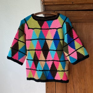 Op de afbeelding: Een kleurrijke trui in vintage-stijl met een geometrisch ruitpatroon. De trui heeft een ronde hals en korte mouwen met zwarte boorden. De ruiten zijn in tinten roze, blauw, groen en bruin, wat zorgt voor een levendig, opvallend ontwerp.