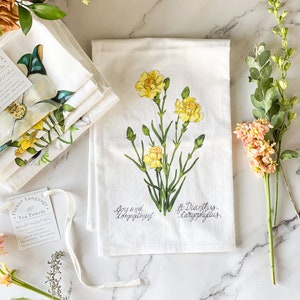Op de afbeelding: Een witte theedoek met een gele bloemenillustratie en de tekst "Vreugde en toewijding" en "Diantus Caryophyllus". De theedoek maakt deel uit van een set theedoeken met bloementaal.