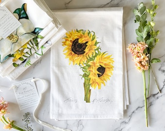 Op de afbeelding: Twee witte theedoeken met een bloemenmotief. Een theedoek heeft een boeket zonnebloemen met de tekst "Having a Blast" in zwarte cursieve letters. De andere theedoek heeft een witte magnolia-bloem met groene bladeren en de tekst "Flower Language Tea Towels" in zwarte cursieve letters.