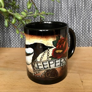 May include: Black ceramic mug featuring a vintage-style illustration of a magpie, a red rose, and a banner that reads "Keepers".