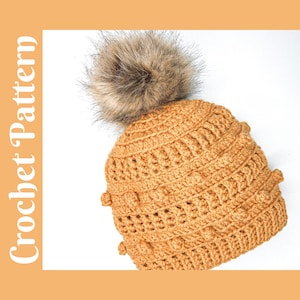 May include: A crocheted beanie hat in a warm brown color with a fluffy brown pom pom on top. The hat has a textured stitch pattern and a slouchy fit.