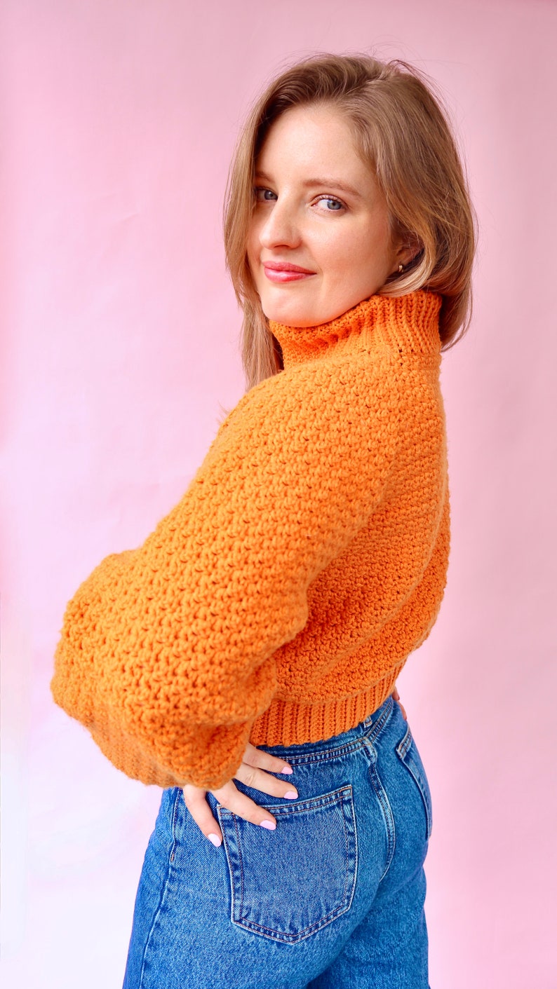 May include: A woman wearing a bright orange turtleneck sweater with a crocheted texture and blue jeans.