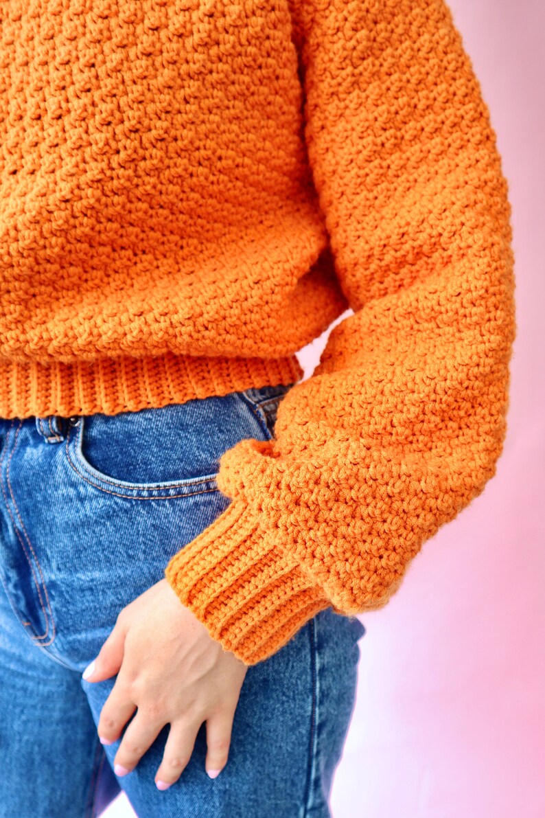 May include: A close-up of a crocheted orange sweater with long sleeves and a ribbed hem. The sweater is worn over blue jeans.