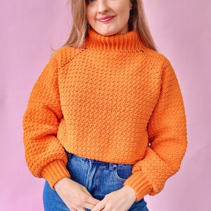 May include: A bright orange crocheted sweater with a high neck and long sleeves. The sweater is cropped and has a loose fit.