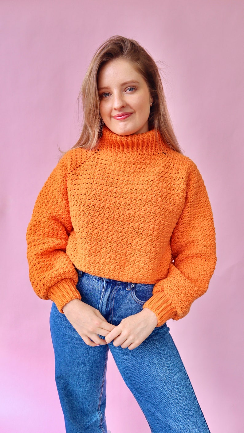 May include: A bright orange crocheted sweater with a high neck and long sleeves. The sweater is cropped and has a loose fit.