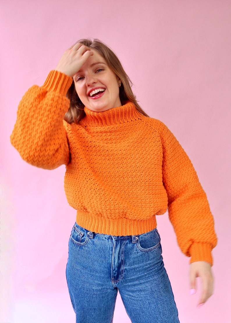 May include: A bright orange, crocheted sweater with a high neck and long sleeves. The sweater has a textured, puffy design and is cropped at the waist. The model is wearing blue jeans.