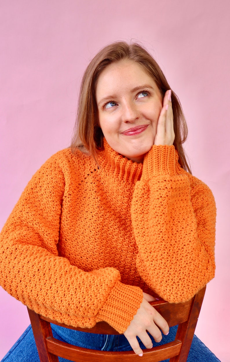 May include: A woman wearing a bright orange crocheted sweater with a high neck and long sleeves. She is sitting on a wooden chair with her hand on the back of the chair. She is looking up and smiling.