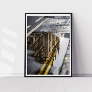 May include: A black and white photograph of a building reflected in a puddle on a city street. The building is tall and has many windows. The street is wet and there are yellow lines painted on it.