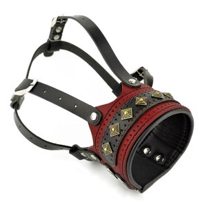 May include: A red leather dog muzzle with black leather straps and gold-toned accents. The muzzle has a decorative design with small squares and a band of gold-toned studs.